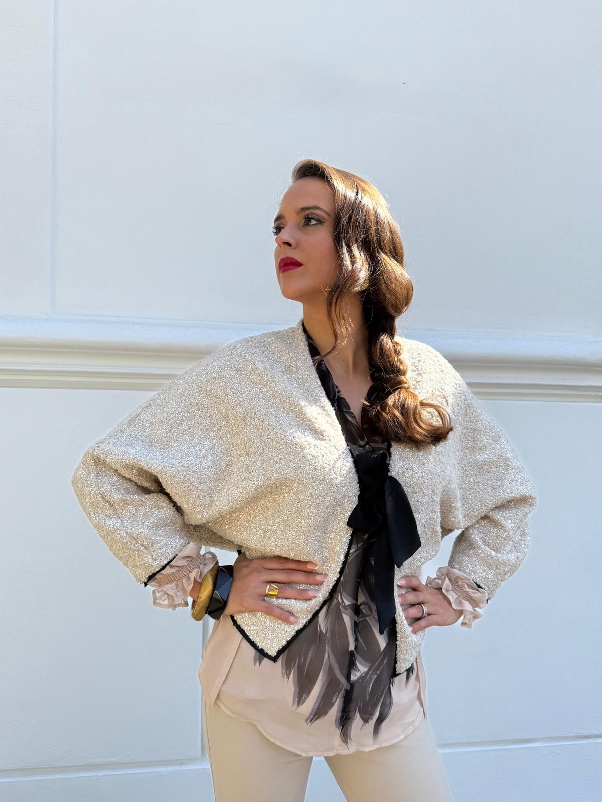 Chaqueta corta de lentejuelas en tono champán con ribete negro y lazada al cuello. Elegante, luminosa y perfecta para completar looks de fiesta o eventos.